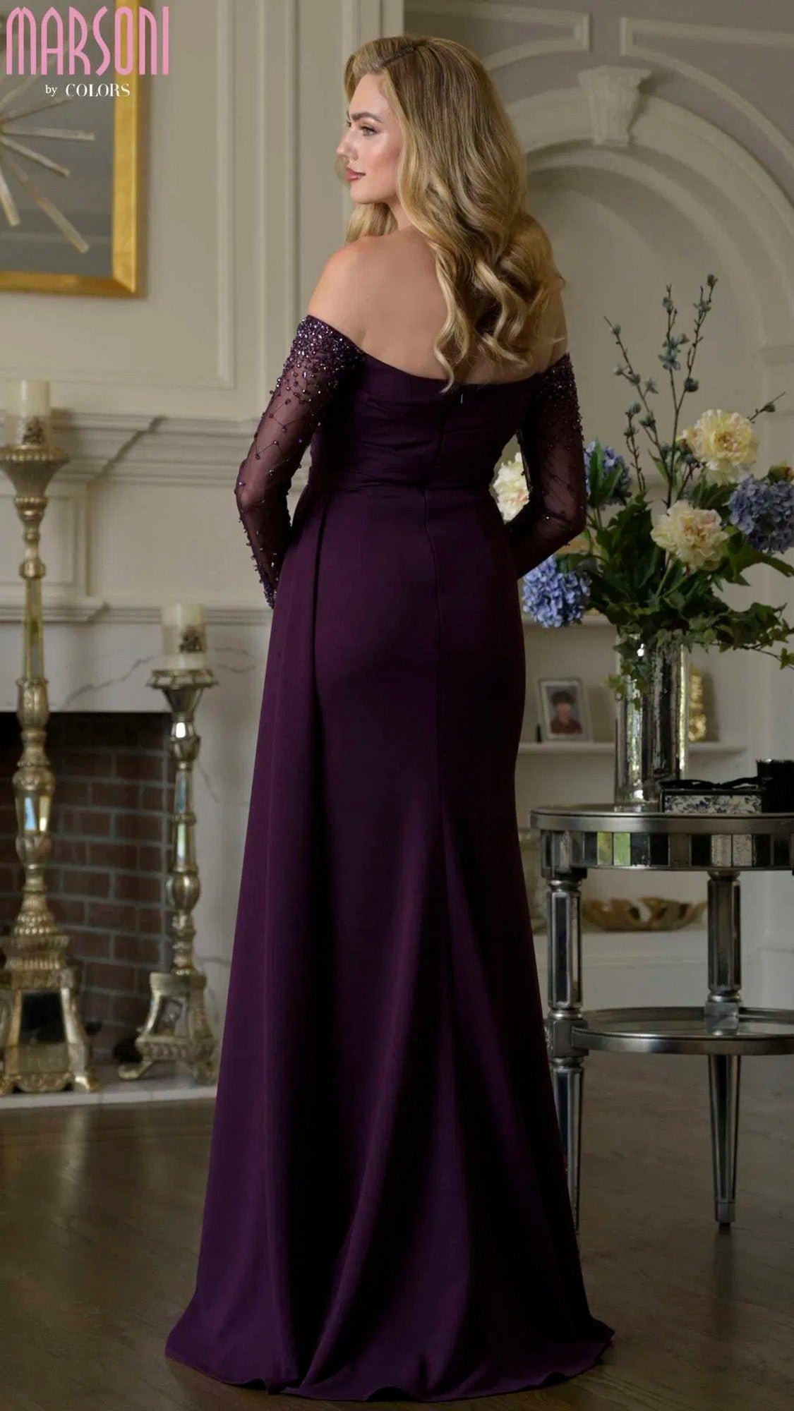 A woman with long, wavy blonde hair wears the Marsoni M344 Long Formal Dress in a deep purple sheath silhouette, standing elegantly in a traditionally decorated room, facing away.